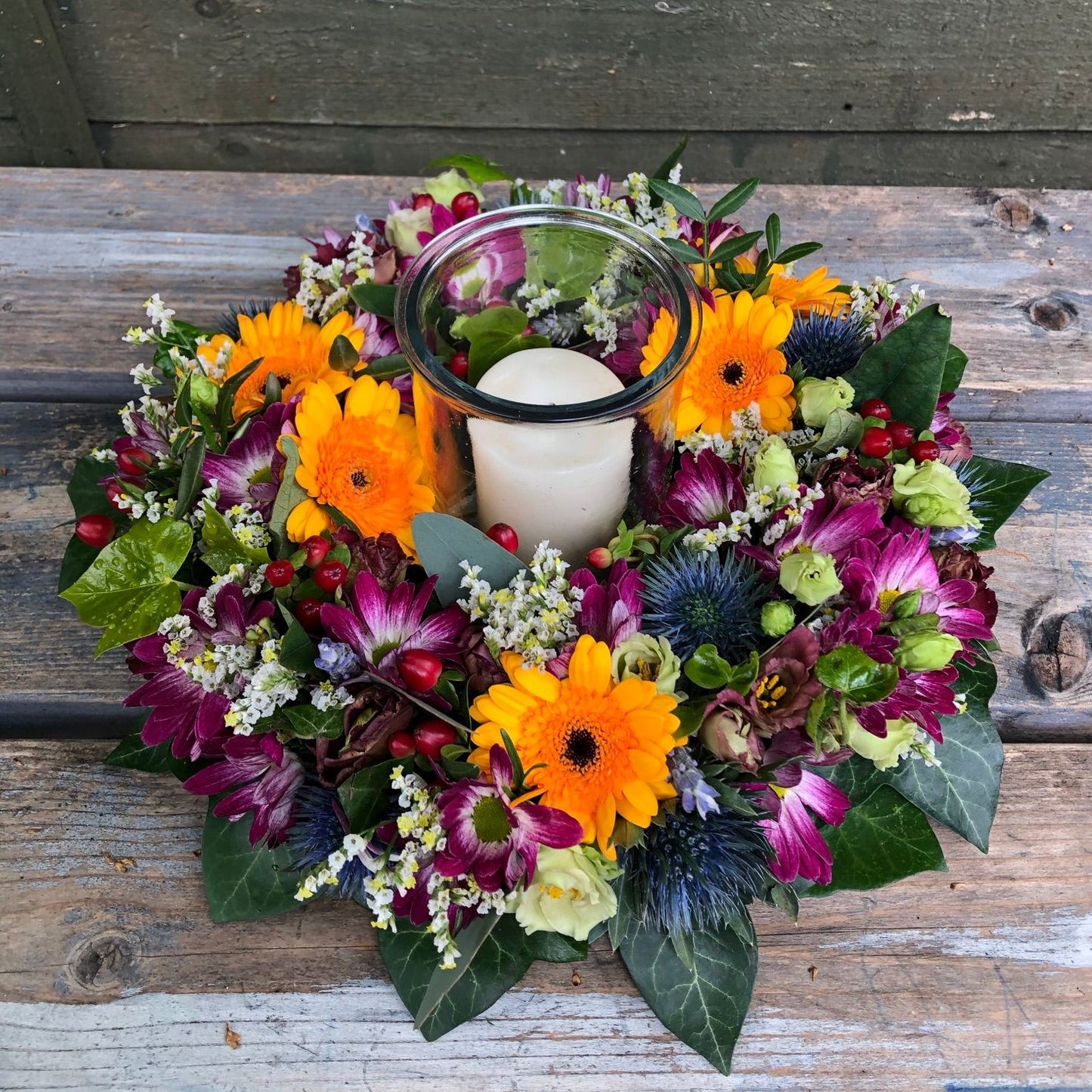 Pictured here is our ' Autumn Table Centre ' which is a fresh flower arrangement with a vase and candle created by our florist. In this arrangement are yellows, purples, blues and whites, flowers including Germinis, Thistles, Chrysanthemums, Berries and more. Due to availability the one we'd create for you may differ slightly to what is pictured but the overall look and style will be the same.