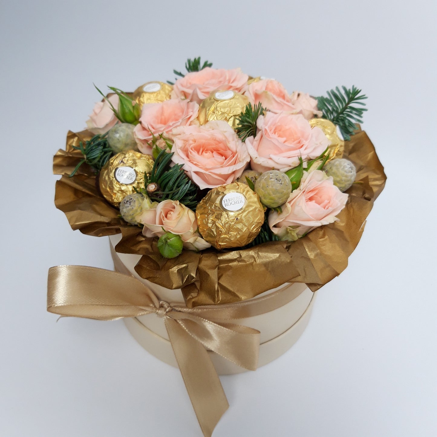 A side view of our cream 'Christmas Sweet Treat Hatbox', filled with pale pink spray roses, brunia, pine, and luxury Ferrero Rocher chocolates. The hatbox is finished with a tied gold ribbon.